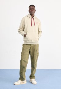 Sudadera beige con un bolsillo frontal tipo bolsa y cordones rojos, combinada con pantalones cargo verde olivo que cuentan con múltiples bolsillos laterales. Zapatillas blancas.