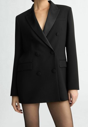 Femme portant une robe blazer noire croisée avec des revers en satin et des collants noirs transparents sur un fond uni.