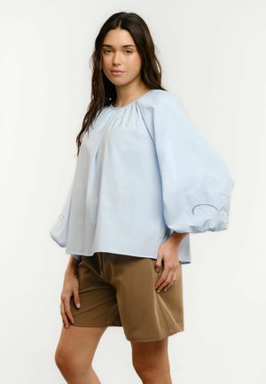 Young woman with long dark hair wears a loose light blue blouse with balloon sleeves and tan knee-length shorts, standing against plain background.