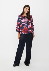 Blumenbluse aus Navy-, Rosa- und Rotdruck mit 3/4 Glockenärmeln; kombiniert mit lockeren, navyblauen Hosen und schwarzen Schuhen.