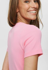 Tânără zâmbitoare care se uită peste umăr, purtând un tricou roz cu mânecă scurtă și textură ribbed, pe un fundal simplu.