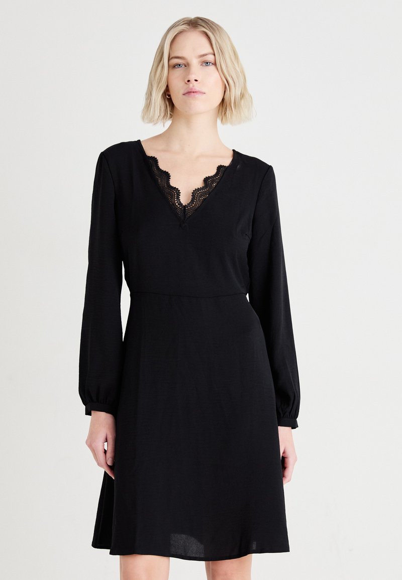 Μαύρο φόρεμα με μακρυά μανίκια και V-σχήμα λαιμόκοψη, διακοσμημένο με δαντέλα. Λείο ύφασμα με εφαρμοστή κορσέ και ρέουσα φούστα.