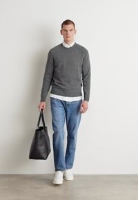 Maglione grigio a maglia, camicia bianca con colletto, jeans azzurri chiari e scarpe da ginnastica bianche. Borsa tote nera in mano, in piedi contro uno sfondo neutro.