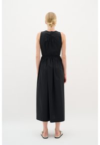 Schwarzes ärmelloses Kleid mit gerafftem Rücken, Taillenknoten und knöchellangem Saum. Glattes Material, minimalistisches Design, ohne Muster oder Akzente.