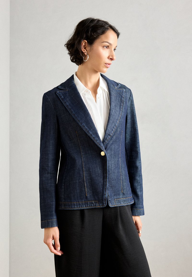 Mørk denimblazer med en enkelt gullknapp, hakkede slag og kontraststing. Bært over en hvit skjorte og svarte bukser.