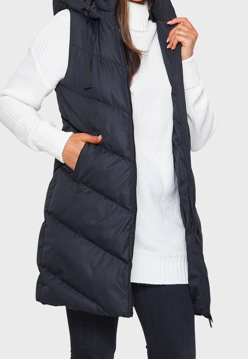 Gilet matelassé noir avec capuche, doté d'une fermeture éclair à l'avant, d'un design en chevron et de deux poches latérales. Porté par-dessus un pull en tricot blanc.