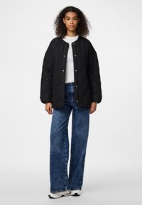Giacca trapuntata nera con chiusure a pressione, scollatura rotonda, maniche lunghe e vestibilità ampia, abbinata a jeans in denim blu a gamba larga e sneakers.