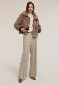 Giubbotto bomber marrone peloso con cerniera nera, maglione lavorato a maglia color crema, pantaloni beige a pieghe e décolleté marroni con punta. Borsa clutch marrone in mano.