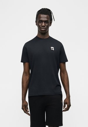 Mężczyzna z plecionymi włosami nosi czarną koszulkę z małym białym logo na piersi oraz czarne szorty na jasnym, jednolitym tle.