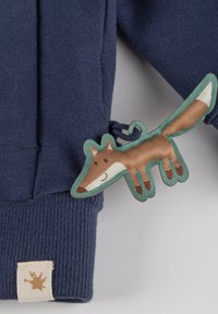 Marineblaues Sweatshirt mit gerippten Bündchen und einer Stofffüchse-Applikation in Braun und Creme mit einem türkisfarbenen Rand, die mit einer grünen Schlaufe befestigt ist.
