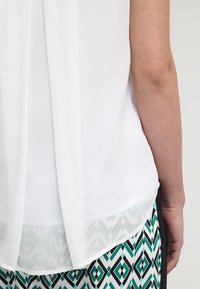 Braccio e vita di una persona che indossa una blusa bianca a strati e una gonna con motivo geometrico verde, nera e bianca.