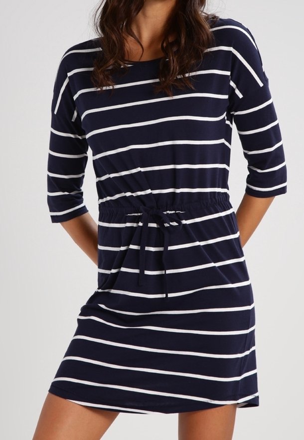 Femme portant une robe bleu marine avec des rayures blanches horizontales, manches trois-quarts, et une ceinture nouée à la taille, debout devant un fond uni.