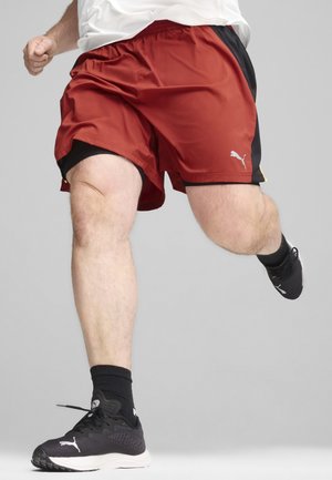 Pantalones cortos deportivos rojos con acentos negros, con una cinturilla elástica y un logo de Puma. Combinados con zapatos deportivos negros y calcetines.