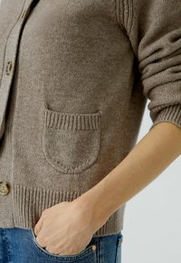 Brauner Strickcardigan mit Knopfverschluss, ausgestattet mit gerippten Bündchen und einer aufgesetzten Tasche auf der linken Seite. Weicher Stoff und leichtes Design.