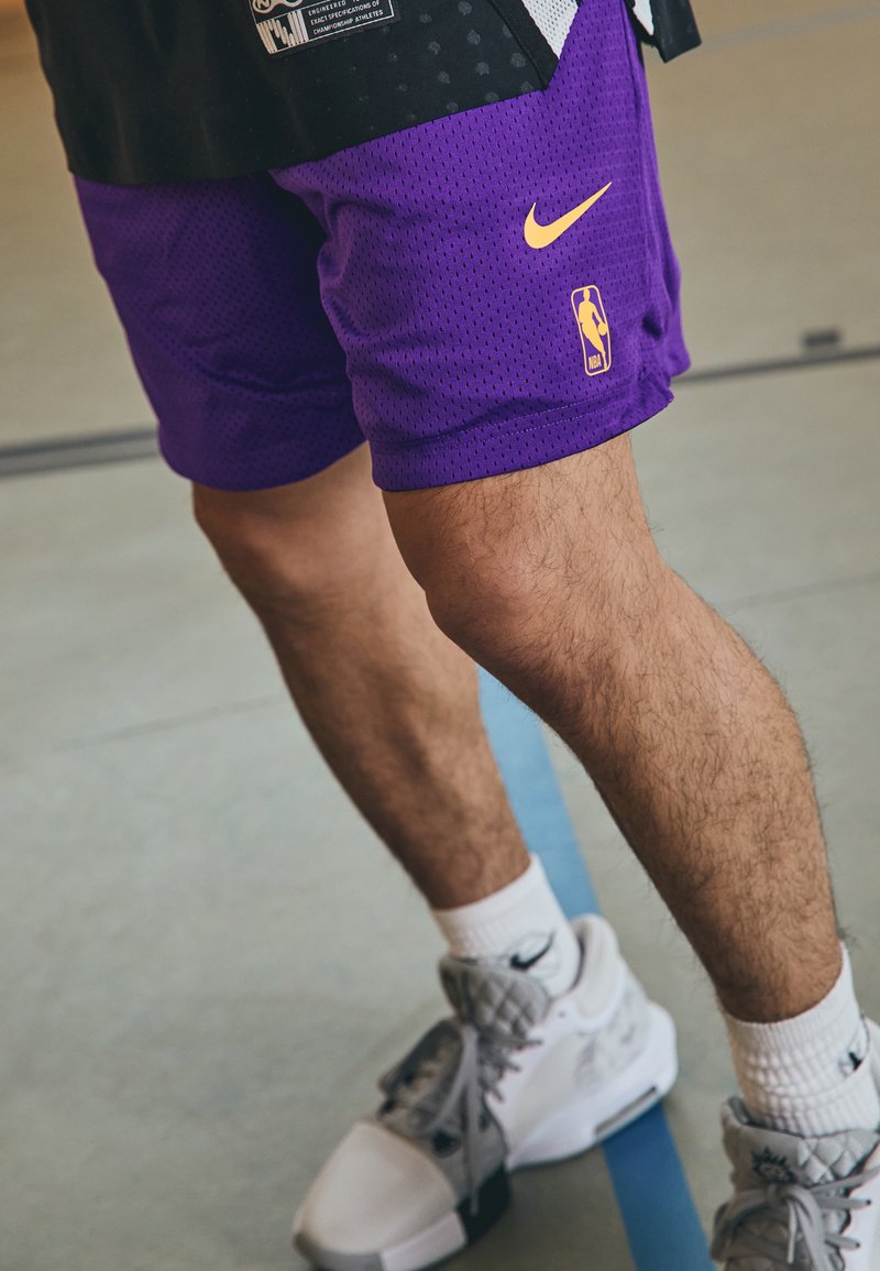 Vijolične košarkarske kratke hlače z mrežasto teksturo, opremljene z rumenim Nikeovim logotipom in logotipom NBA. Združene s sivimi in belimi supergami.