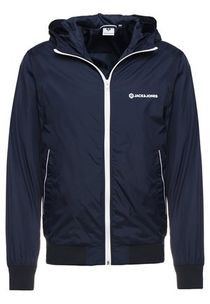 Lekka granatowa kurtka z kapturem, białym zamkiem błyskawicznym, białym logo na piersi, bocznymi kieszeniami z białą lamówką oraz czarnymi ściągaczami przy rękawach i u dołu.