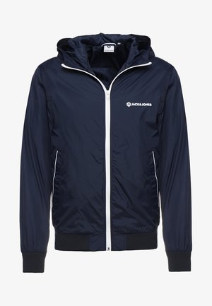 Chaqueta ligera azul marino con capucha, cremallera blanca, logo blanco en el pecho, bolsillos laterales con ribete blanco y puños y dobladillo negros acanalados.