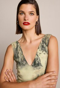 Grön och kräm-mönstrad klänning med djup V-ringning, ärmlös design och en texturerad tyg; modellen har kort hår och guld hoopsörhängen.
