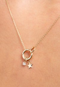 Goudkleurige ketting met twee aan elkaar gekoppelde ringen, met een hangende heldere edelsteen en een stervormige bedel. De ketting heeft getextureerde schakels.