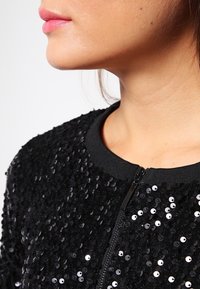 Kvinnas nedersta ansikte och nacke som bär en svart paljettzip-up topp med rund halsringning mot en enfärgad bakgrund.