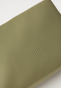 Zelený vodotesný obal vyrobený z matného materiálu, s embosovaným názvom značky "RAINS" a hladkým, elegantným dizajnom.