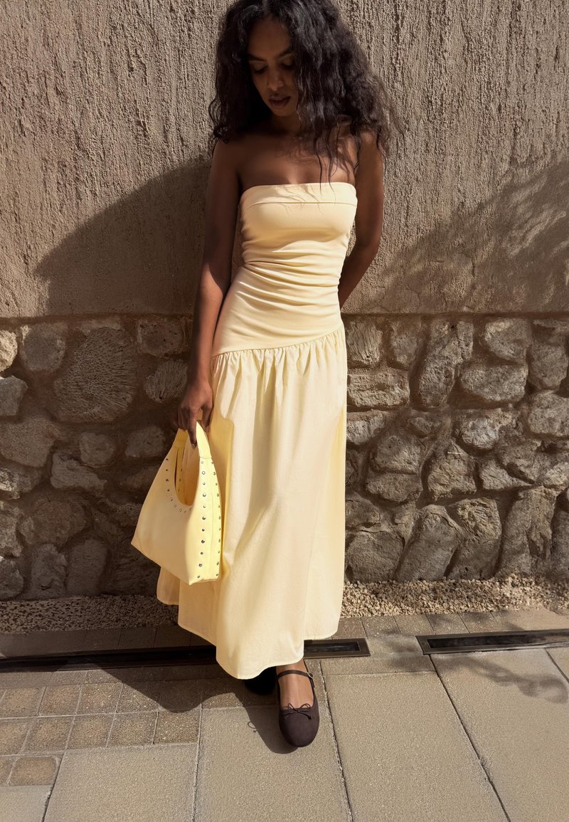 Femme aux longs cheveux bouclés portant une robe jaune sans bretelles, tenant un sac à main jaune, debout contre un mur en pierre et stuc sous la lumière du soleil.