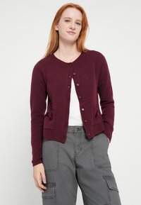 Maglione bordeaux a maniche lunghe, con scollo rotondo, bottoni frontali e due tasche applicate con fiocchi, abbinato a pantaloni cargo grigi.