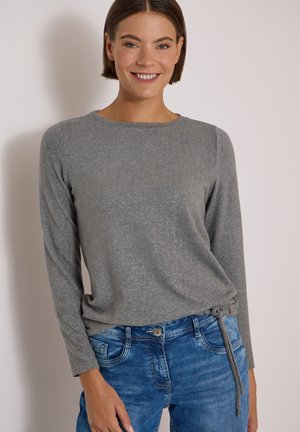Jeune femme souriante, portant un haut gris à manches longues avec des liens sur les côtés et un jean bleu, debout devant un fond clair uni.