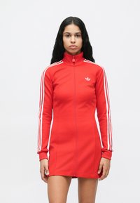 Sieviete, tērpusies sarkanā Adidas zīmogotā kleitā ar baltām svītrām uz piedurknēm un logotipu uz krūškurvja, stāv uz vienkrāsaina baltā fona.