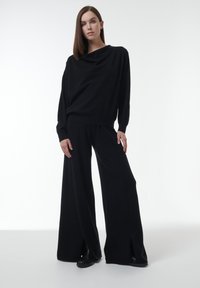Zwarte gebreide trui met een gedrapeerde halslijn en lange mouwen, gecombineerd met zwarte broek met wijde pijpen en voorsluitingen. Soepele textuur.