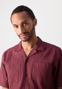 Tamsiai raudona trumpomis rankovėmis marškiniai su tekstūruotu raštu, dviem priekinėmis kišenėmis ir apykakle. Yra mygtukų uždarymas ir laisvas siluetas.