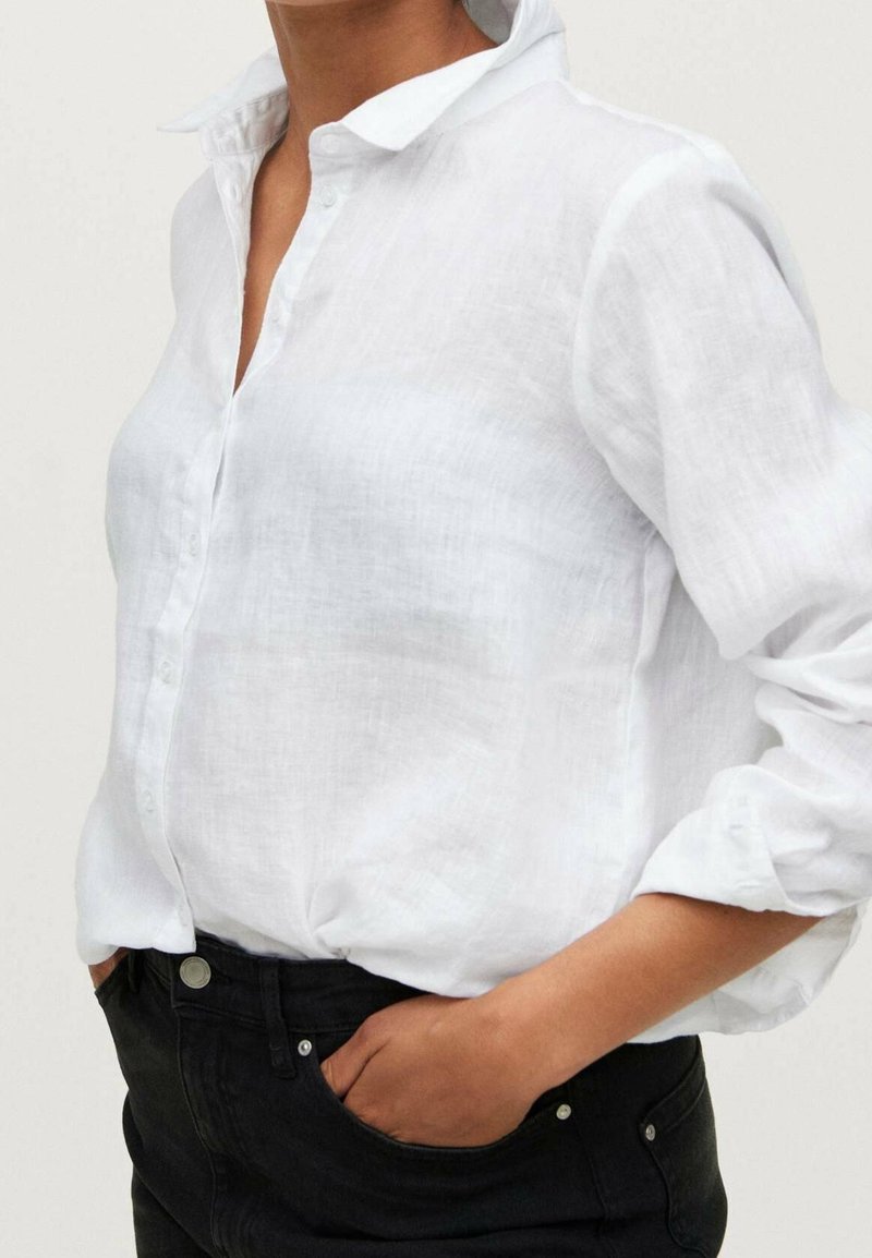 Camicia bianca in lino con colletto e maniche arrotolate, abbinata a jeans neri. Vestibilità rilassata, tessuto leggero.