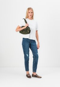 Žena v bielom tričku s krátkym rukávom, v modrých džínsoch s poškodeným vzhľadom, s topánkami s leopardím vzorom, drží zelenú kabelku cez rameno na bielom pozadí.