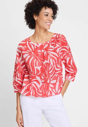 Red blouse with white tropical leaf pattern, boat neckline, gathered detail at shoulders, and three-quarter sleeves. Paired with white pants.