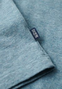 Light blue fabric with a soft texture and a heathered pattern. Features a small dark blue tag with white text sewn into the fabric.