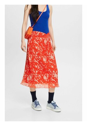 Femme portant un débardeur bleu, une jupe midi rouge à motifs, des chaussettes noires, des baskets grises et un sac bandoulière orange, debout sur un fond blanc.