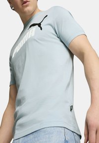 Camisa de manga corta azul claro hecha de mezcla de algodón, con un prominente logo blanco de "PUMA" y un emblema de gato negro en el pecho.