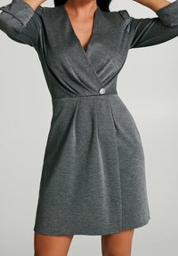 Szara sukienka o kroju kopertowym, uszyta z miękkiego, teksturowanego materiału. Posiada głęboki dekolt w kształcie litery V, marszczone detale, centralny guzik oraz dopasowaną talię z plisami.