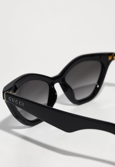 Occhiali da sole neri a forma di occhio di gatto con lenti scure, caratterizzati da un logo con accenti dorati sulla stanghetta. Materiale in plastica liscia con finitura lucida.