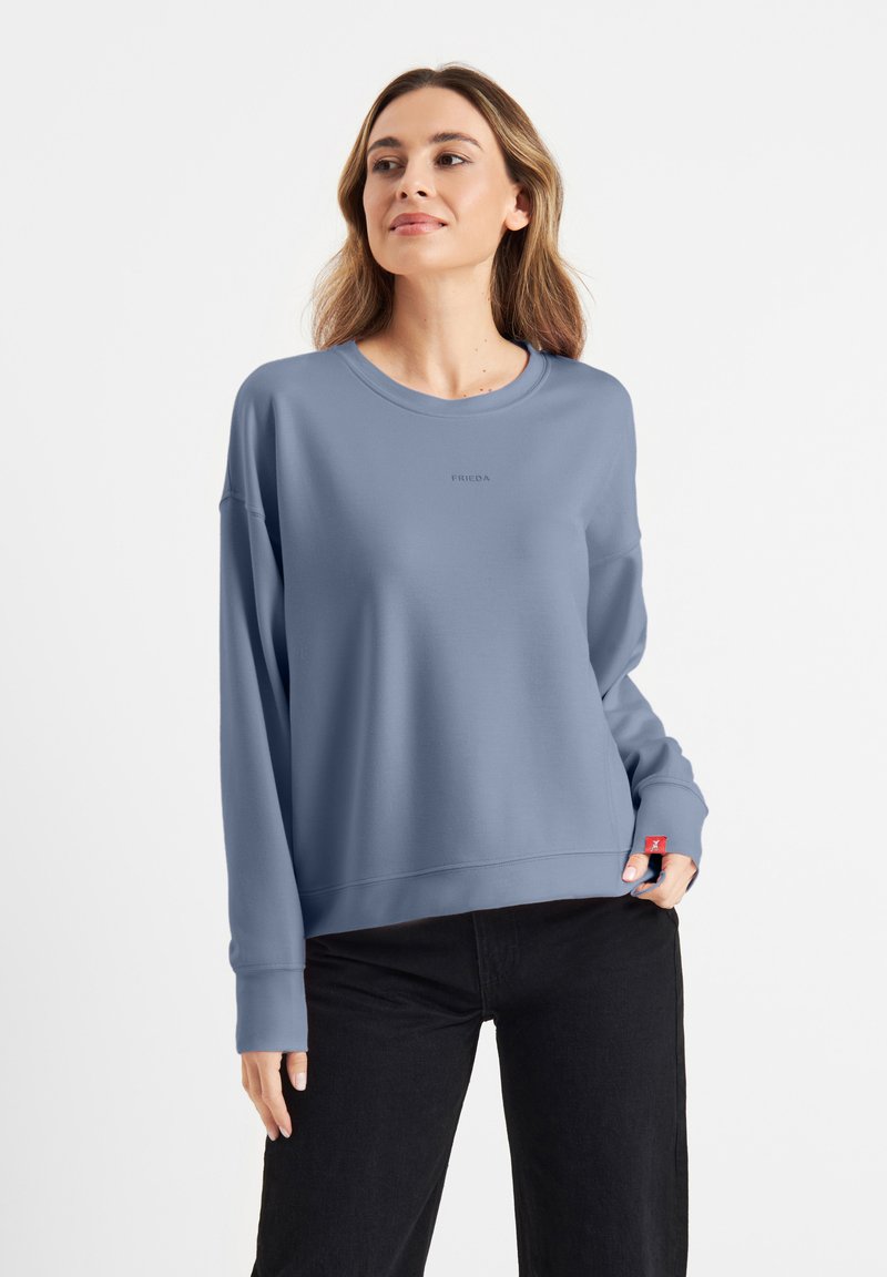 Jasnoniebieski sweatshirt z okrągłym dekoltem, luźnym krojem i długimi rękawami. Posiada małe logo na klatce piersiowej oraz czerwony akcent na dolnym brzegu.