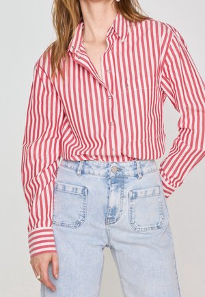 Femme portant une chemise boutonnée à rayures verticales rouges et blanches rentrée dans un jean bleu clair taille haute avec des poches plaquées à l'avant.