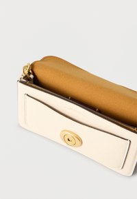 Μικρή κρεμ τσάντα από δέρμα με στρογγυλό χρυσό κούμπωμα και καφέ σουέντ εσωτερικό, πάνω σε απλό ανοιχτό φόντο.