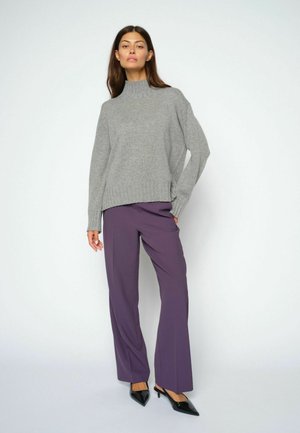 Donna che indossa un maglione grigio a collo alto, pantaloni viola a gamba larga e tacchi neri a punta, in piedi con una mano in tasca.