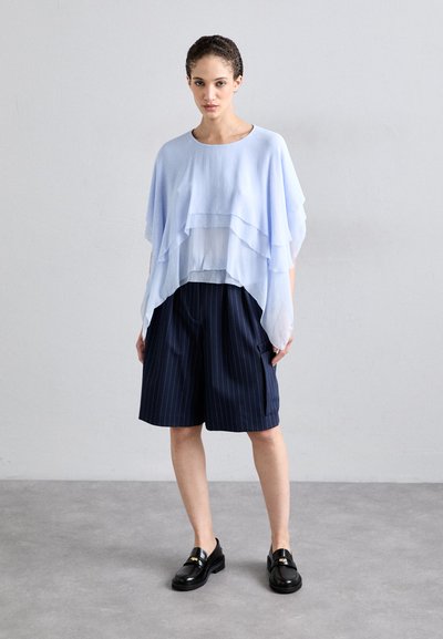 Lichtblauw gelaagd topje, met vloeiende stof, gecombineerd met marineblauwe gestreepte shorts. Model draagt zwarte instappers en staat op een neutrale grijze achtergrond.