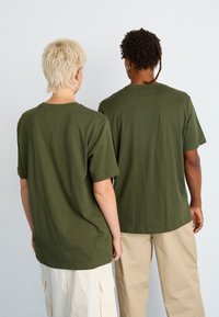 Dwie osoby stojące plecami do kamery, ubrane w dopasowane zielone koszulki, jedna z krótkimi jasnymi włosami i białymi spodniami, a druga z kręconymi ciemnymi włosami i beżowymi spodniami.