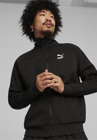 Svart zip-up jacka med en texturerad yta, som har en liten vit Puma-logga på bröstet och kontrasterande sidostråk.