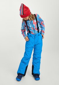 Pantalons de neige bleu vif avec des bretelles noires ajustables, associés à un bonnet rouge à rayures et un haut à manches longues à motifs multicolores.