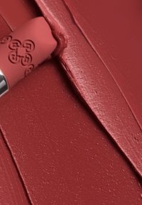 Rossetto in una tonalità rossa calda opaca con una texture liscia, parzialmente applicato, che mostra un cappuccio inciso con un logo argentato e una finitura cremosa.