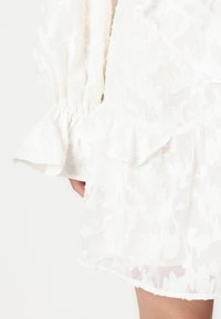 Λευκό φόρεμα με υφασμάτινο λουλουδάτο σχέδιο, ριγωμένα μανίκια και τελείωμα. Κατασκευασμένο από ελαφρύ ύφασμα, με μαλακή, διάφανη όψη.