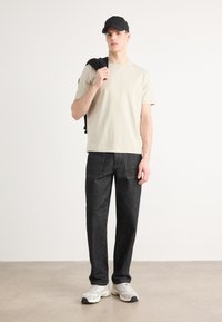 Νεαρός άνδρας που φοράει μαύρο καπέλο, μπεζ μπλουζάκι, μαύρο τζιν και λευκά αθλητικά παπούτσια, κρατώντας ένα μαύρο μπουφάν στον ώμο του, στέκεται σε ουδέτερο δάπεδο.